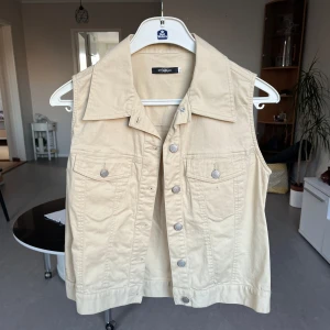 Beige jeansväst  - Snygg beige jeansväst med knappar framtill. Perfekt för att lägga till en cool touch till din outfit. Västen har en klassisk krage och två bröstfickor med knappdetaljer.