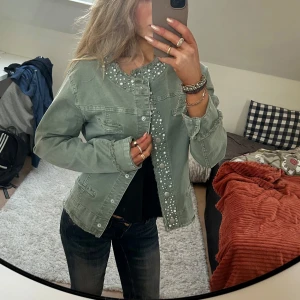 Grön jeansjacka med pärlor - Snygg grön jeansjacka med pärldetaljer längs kragen och knäppningen. Jackan har en normal passform och långa ärmar. Uppskattar storlek    S/M. Perfekt nu till våren!