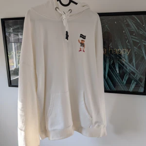 Vit hoodie från Palm Angels - Säljer en vit hoodie från Palm Angels med svart text på framsidan och en söt nalle med flagga på baksidan. Hoodien har en stor ficka framtill och justerbar dragsko i huvan. Perfekt för en avslappnad stil!