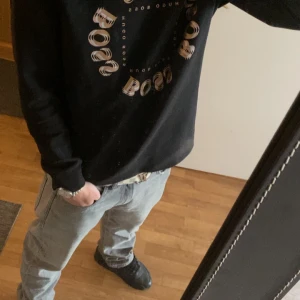 Svart tröja från Hugo Boss - Säljer en svart tröja från Hugo Boss med ett cirkulärt mönster av märkesnamnet på bröstet. Tröjan har långa ärmar och en klassisk passform. Perfekt för en stilren look.