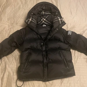 Svart dunjacka ifrån burberry - Säljer en svart dunjacka ifrån burberry rutigt foder i huvan.Jackan har dragkedja och knappar framtill samt långa ärmar. Perfekt för kalla dagar med sin quiltade design.