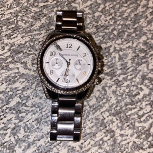 Silverfärgad klocka från Michael Kors - Snygg och stilren klocka från Michael Kors i silverfärg. Urtavlan är vit med tydliga siffror och kronograffunktion. Armbandet är i rostfritt stål och har en klassisk design. Perfekt för den som vill ha en elegant accessoar.