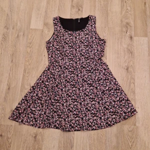 Blommig ärmlös klänning - Söt ärmlös klänning med ett blommigt mönster i rosa och lila på en svart bakgrund. Klänningen har en dragkedja i ryggen och en lätt utställd kjol som ger en fin siluett.