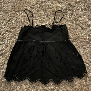 Svart  topp från Zara - Säljer en svart topp från Zara med tunna axelband och broderade detaljer. Toppen har en fin knytning i ryggen och en vågig kant nedtill. Perfekt för varma dagar eller som ett lager under en jacka.