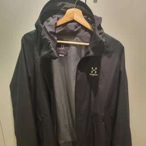 Svart jacka från Haglöfs - Svart jacka från Haglöfs med justerbar huva och dragkedja framtill. Jackan har en stilren design med Haglöfs logga på bröstet. 