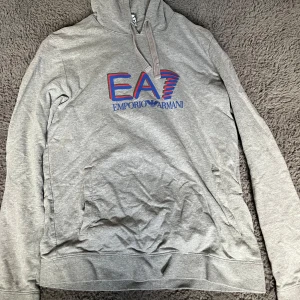 Grå hoodie från Emporio Armani - Säljer en stilren grå hoodie från Emporio Armani med EA7-logga i blått och rött på framsidan. Tröjan har en klassisk huva med dragsko och en praktisk magficka. Perfekt för en avslappnad look.