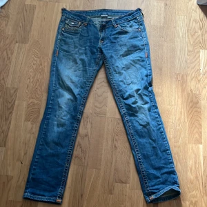 Blå jeans från True Religion - Snygga blå jeans från True Religion med ikonisk broderad detalj på bakfickorna. Jeansen har en klassisk femficksdesign och orangea sömmar som ger en cool kontrast. Perfekta för en avslappnad stil. Ena hämpan trasig går dock att laga. Innebenslängden är 78,5 cm. Midjemått 45 cm.