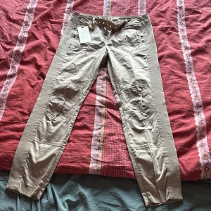 Beige byxor från Bomboogie - Snygga beige byxor från Bomboogie med dragkedjor och knappar. Byxorna har en cool design med flera fickor och ett bälte i midjan. Perfekta för en avslappnad stil.