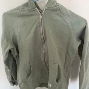Grön hoodie med dragkedja - Säljer en stilren grön hoodie med dragkedja framtill. Tröjan har en praktisk känguruficka och en bekväm huva. Perfekt för en avslappnad look. JAG TROR DEN ÄR XS FÖR DEN KÄNNS SOM DE.