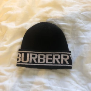 Svart mössa från Burberry - Snygg svart mössa från Burberry med vit logotyp runt kanten. Perfekt för att hålla sig varm och stilren under kyliga dagar. 169 kr endast 18,19,20 mars sedan går den upp till ordinarie pris som är 239 kr.