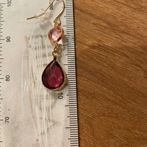 Guldörhängen med rosa stenar - Eleganta guldörhängen med två rosa stenar i olika nyanser. Den övre stenen är rund och ljusrosa, medan den nedre är droppformad och mörkare rosa. Perfekta för att ge en färgklick till din outfit. Gjord av nickel. Har aldrig använt 💞