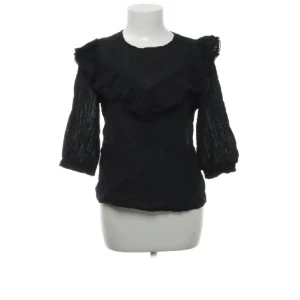 Svart blus med volangdetaljer - Snygg svart blus med volangdetaljer på bröstet och axlarna. Blusen har trekvartsärmar och en rund halsringning. Perfekt för en stilren look.