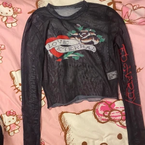 Svart Ed Hardy långärmad topp - Säljer en cool svart långärmad topp från Ed Hardy med tryck av ett hjärta och texten 'Love Eternal'. Toppen har ett unikt mönster och är perfekt för dig som gillar edgy stil. Den har även Ed Hardy-loggan i rött på ärmen.