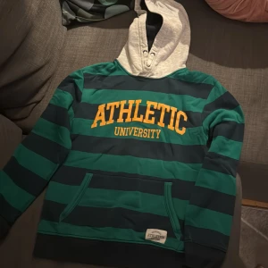 Randig hoodie Athletic University - Snygg hoodie med breda gröna och mörkblå ränder, stor gul text 'ATHLETIC UNIVERSITY' på bröstet och ljusgrå huva. Praktisk magficka framtill och sportig vibe, perfekt för chill dagar eller häng med kompisar.