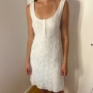 Vit spetsklänning med knappar - Superfin vit kortklänning i spets med ärmlös design och rundad urringning. Klänningen har dekorativa knappar framtill!