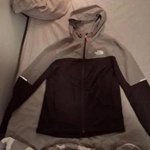 Svart och grå vindjacka från The North Face - Säljer en snygg svart och grå vindjacka från The North Face i herrmodell, storlek S. Jackan har huva, hel dragkedja och logga på bröstet och ryggen. Perfekt för blåsiga dagar och har en clean, sportig look. Materialet känns lätt och smidigt. Samt en liten skada vid luvan