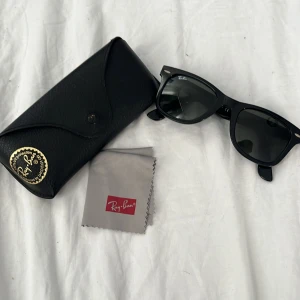 Ray-Ban Wayfarer svarta solglasögon - Klassiska Ray-Ban Wayfarer solglasögon i svart med ikonisk fyrkantig form. Snygg och tidlös design med mörka glas och diskret Ray-Ban-logga på linsen. Kommer med originalfodral och putsduk. Perfekta för att lyfta din stil i sommar! Passar till vuxen/ ungdom