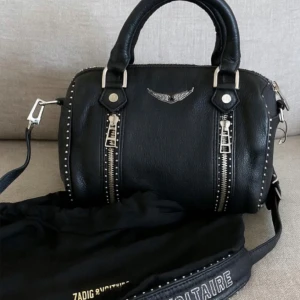 Zadig & Voltaire väska - Zadig & Voltaire sunny bag med silvriga detaljer, dubbla dragkedjor framtill och nitar längs kanterna. Väskan har både handtag och axelrem med logga, samt en snygg vinge i metall framtill. Dustbag ingår och en tagg finns kvar. 