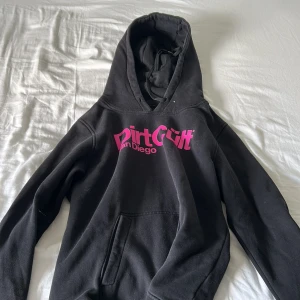 Svart hoodie Dirt Cult San Diego - Svart hoodie från Dirt Cult San Diego med stor rosa logga på bröstet. Klassisk modell med huva och känguruficka framtill. Perfekt för dig som gillar streetwear och vill sticka ut med en fet tryckdetalj.