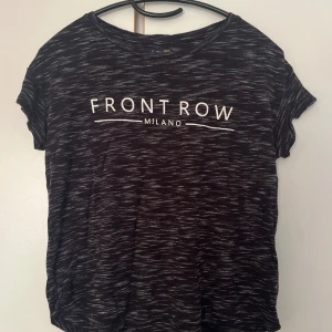 Svartvit t-shirt Crocker Front Row - Svart t-shirt från Crocker med vit text 'Front Row Milano' på bröstet. T-shirten har korta ärmar, rund hals och ett melerat mönster i svart och vitt. Skön passform och tillverkad i mjukt bomullsmaterial. Perfekt för en chill och stilren look.