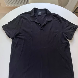 Svart pikétröja från Hugo Boss storlek 3XL - Klassisk svart pikétröja från Hugo Boss i mjuk pima bomull. Tröjan har korta ärmar, krage med knappslå och diskret broderad logga på bröstet. Perfekt för en clean och stilren look.