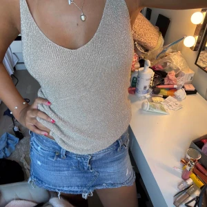 Glittrig beige linnetopp - Säljer en glittrig beige/guldigt linnetopp med smala axelband och djup urringning. Toppen har en figurnära passform och är perfekt för dig som vill sticka ut lite extra. Materialet är mjukt och skimrande, vilket ger en snygg effekt i ljuset.