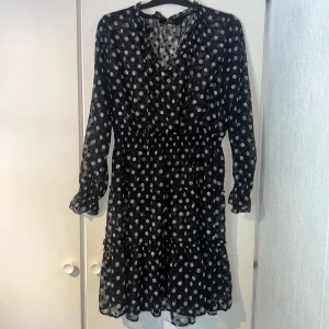Svart prickig kortklänning - Supersöt svart kortklänning från Capri Collection med vita prickar. Klänningen har v-ringning, långa ärmar med resår vid ärmslut och volangdetaljer nertill. Tunt, transparent material som ger en luftig känsla. Perfekt för dig som gillar lekfulla mönster! Inköp för ca 800kr. Fåtal använd. Passar S-XL