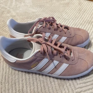 Adidas Gazelle i rosa mocka - Snygga Adidas Gazelle sneakers i ljusrosa mocka med klassiska vita ränder och vit häl. Skorna har platt sula, rund tå och snörning. Perfekta för dig som vill ha en stilren och trendig look med en touch av retro. Använd 1 gång! 