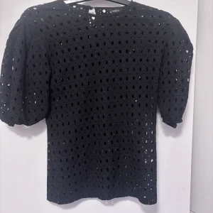 Svart blus med puffärm från Lindex XS - Svart blus från Lindex i storlek XS med coola puffärmar och genombrutet hålmönster över hela toppen. Blusen har rund halsringning och knäppning bak i nacken. Perfekt för dig som gillar unika detaljer och vill sticka ut lite extra.