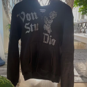 Svart Vonstudio hoodie med strass - Svart hoodie från Vonstudio med dragkedja och huva. Framsidan har stora strasstenar som bildar texten 'Von Studio' och en cool ängelgrafik. Mjuk insida och känguruficka framtill. Perfekt för dig som gillar streetwear med bling. Hoodien passar på både M och S där jag har M och den sitter väldigt mot huden så. 