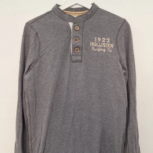 Grå långärmad tröja Hollister  - Grå långärmad tröja från Hollister med broderad text '1922 Hollister Surfing Co.' på bröstet. Tröjan har tre knappar framtill och en ribbad krage. Skön och avslappnad passform, perfekt till jeans eller shorts.