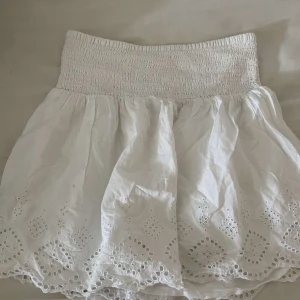 Vit broderad kortkjol från Gina Tricot - Supersöt vit kortkjol från Gina Tricot med bred smock i midjan och broderade detaljer längst ner. Kjolen är i bomull och har en luftig känsla med fina hålbroderier och vågig kant. Perfekt för varma dagar och ger en fräsch sommarvibe.