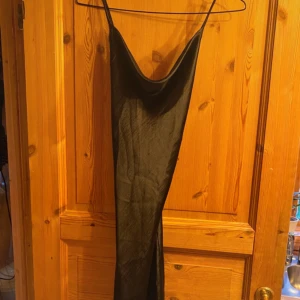 Svart långklänning i satin med slits - Svart långklänning i glansigt satinliknande material med tunna axelband och snygg draperad urringning. Klänningen har en slits på sidan och smal passform, perfekt för dig som gillar en elegant och stilren look. Snörning därbak. Xs/S är storleken beroende på hur hårt man knyter snören i ryggen.