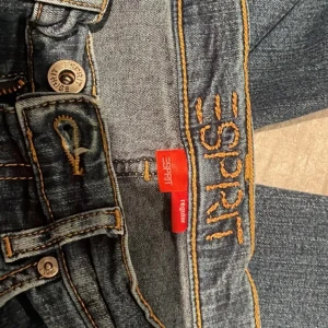 Blå bootcut jeans från Esprit - Snygga blå jeans från Esprit med bootcut passform och coola broderade detaljer på bakfickorna. Klassisk femficksmodell med orange kontrastsömmar och regular midja. Omsydda för en mer trending och unik look. Billigt, snyggt och miljövänligt! Storlek 34/S