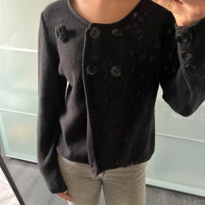 Svart stickad kofta med knappar - Säljer en svart stickad kofta med asymmetrisk knäppning och stora svarta knappar framtill. Koftan har lång ärm och en dekorativ knutdetalj vid axeln. Perfekt för lager-på-lager och enkel att styla till olika outfits.
