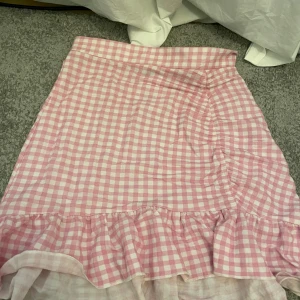 Rutig rosa minikjol med volang - Supersöt kortkjol i rosa och vitt med rutigt mönster. Kjolen har en volangkant nertill och lätt rynkade detaljer på ena sidan. Perfekt för dig som gillar en gullig och somrig stil!
