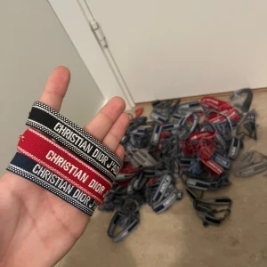 3 stycken Christian Dior J'Adior armband i tyg - Säljer flera Christian Dior J'Adior armband i tyg med broderad logga. Finns i färgerna svart, röd, blå och grå med matchande tofsar. Perfekt accessoar för att lyfta din outfit lyxig touch och cool streetstyle-vibe.