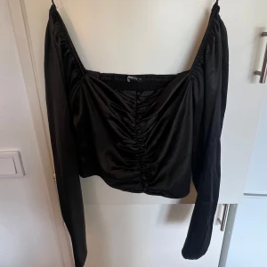 Svart fest-topp med knappar - Toppen har knappar och rynkade detaljer framtill samt en smockad rygg för extra komfort. Gjord i glansigt material som ger en lyxig känsla och passar perfekt till fest🌻