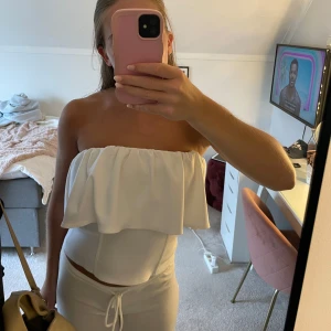 Vit bandeau tubtopp med volang - Säljer en vit bandeau tubtopp med bred volang upptill. Toppen är axelbandslös och har en figurnära passform som sitter snyggt över kroppen. Perfekt för dig som vill ha en clean och stilren look med lite extra detalj upptill.