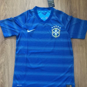 Brasilien 2014 Borta tröja  - Snygg blå Brasilien fotbollströja från Nike med CBF-emblem och fem stjärnor på bröstet. Tröjan har korta ärmar, V-ringning och är tillverkad i Dri-FIT-material som andas. Mönstret består av små prickar i olika blå nyanser för en modern look.        Brasilien 2014 Away Kit, Storlek Herr S med prislappen fortfarande kvar.