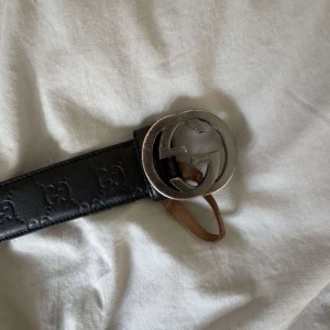 Svart Gucci skärp med GG-spänne - Säljer ett svart skärp från Gucci i äkta skinn med präglade GG-loggor längs hela bältet. Stort, rundat silverspänne med klassiska dubbla G. Insidan är brun med tydliga märkningar. Kommer med originalpåse från Gucci. Det har blivit lite rost på bältet annars finns de inga defekter. Bältet är självklart äkta, priset är ej huggen i sten. Tveka inte med att skriva några funderingar!!😃