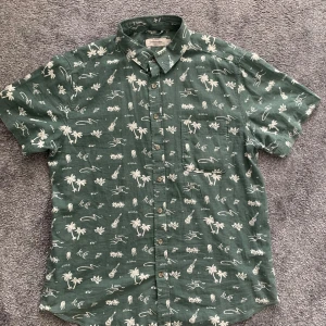 Grön linneskjorta med mönster Dressmann - Grön kortärmad skjorta från Dressmann i linnemix med vita tropiska motiv som palmer, fiskar och gitarrer. Skjortan har klassisk krage, bröstficka och knappar framtill. Perfekt för chill sommardagar när du vill sticka ut lite extra.