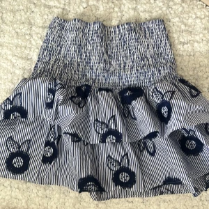 Holly&White Randig volangkjol med broderi - Supersöt kort kjol med blå och vita ränder, volanger och broderade blommor. Resår i midjan för skön passform. Perfekt för dig som gillar en lekfull och trendig stil.