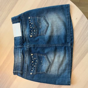 Jeanskjol med stjärnnitar från Simply Chic - Snygg blå jeanskjol från Simply Chic med coola stjärn- och metallnitar på fickorna. Kjolen har klassisk femficksdesign, dubbla silverknappar och en lätt tvättad look. Perfekt för dig som gillar en edgy och trendig stil. Populär så var snabb!