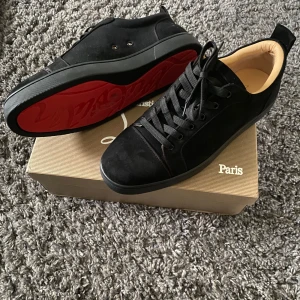 Svarta Loubs - Snygga svarta sneakers från Christian Louboutin i mocka med ikonisk röd sula. Skorna har rund tå, svarta skosnören och diskreta detaljer. Insidan är beige och sulan har Louboutins signatur. Perfekta för dig som vill sticka ut med lyxig streetstyle.