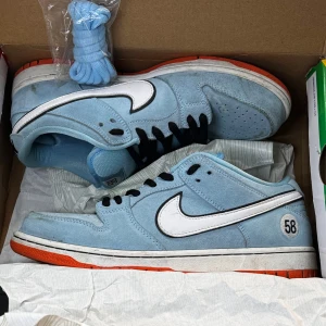 Nike SB Dunk Low GULF - Nike SB Dunk Low gulf 58. Skorna har svarta snören, blåa skosnören medföljer. Finns någon liten fläck på mockan men annars väldigt fina, lite starloss inge heeldrag. Box fås med. 