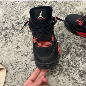 Nike Air Jordan 4 svart/röd sneakers - Säljer ett par Nike Air Jordan 4 sneakers i svart och rött med klassisk Jumpman-logga på plösen och hälen. Skorna har svarta snören, meshdetaljer och röda accenter på sula och snörning. Ovandelen är i mocka och syntet, perfekt för dig som gillar streetstyle.