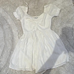 Vit kortklänning från Hollister - Supersöt vit kortklänning från Hollister med broderade detaljer och puffärmar. Klänningen har v-ringning, snörning framtill och är tillverkad i ett lätt bomullstyg. Perfekt för varma dagar och har en romantisk vibe. 