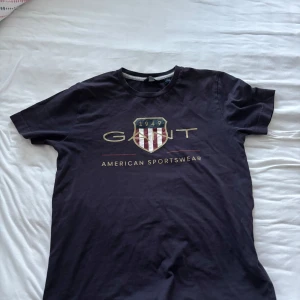 Svart GANT t-shirt med tryck - Svart t-shirt från GANT med stort tryck framtill och klassisk rund hals. T-shirten har korta ärmar och är gjord i mjuk bomull, perfekt för en chill och sportig look. Snygg logga och texten 'American Sportswear' på bröstet.