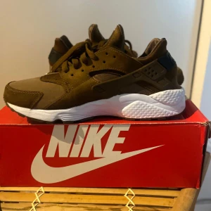 Nike Air Huarache Run PA brun 43 - Nike Air Huarache Run PA i snygg mörkbrun färg med vita detaljer på sulan och svart hälrem. Skorna har en chunky sula, snörning och är tillverkade i mocka och syntet. Perfekta för dig som gillar streetstyle och vill sticka ut med en unik färg på dina sneakers. Obs en söm harr gått upp på ena skon syns på bilden, 